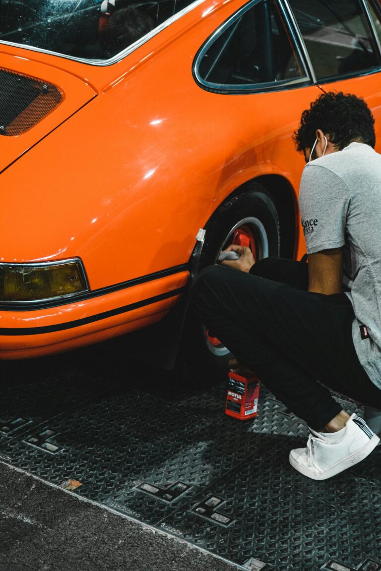 A man carefully cleans the wheel of a classic orange sports car in a garage setting.